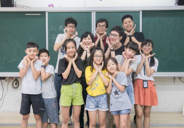 教室內約十五名小學生在綠色黑板前合照，孩子們排成兩排，做出雙手托腮的可愛姿勢，穿著各色校服，表情開朗歡樂，展現班級的溫馨團結氛圍。