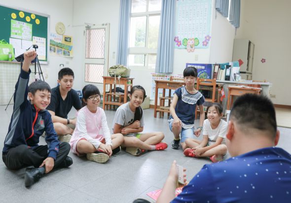 教室內，一群小學生圍坐成圈，進行團體活動。其中一名學生舉起相機拍照，其他學生坐在地板上參與互動。教室背景可見綠色布告欄、書架、窗戶和教室布置，呈現活潑的學習環境。