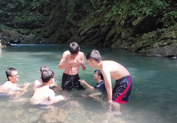 五位中年男性在翠綠色溪水中進行水上活動，周圍被茂密的森林和陡峭的岩壁包圍。其中一人穿著橙色救生衣，其他人則穿著泳褲在齊腰深的清澈溪水中活動。環境優美，是典型的山區溪流探險場景。