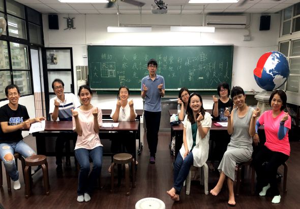 教室內約十位學生圍坐成圈，中央站著一位男性講師。黑板上寫滿數學公式，教室裡懸掛著紅藍色地球儀。學生們穿著休閒，多數人拿著飲料或食物，氛圍輕鬆。這是一場互動式的教學或討論活動。