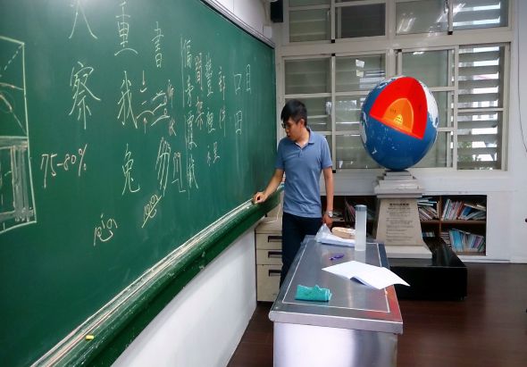 教室內，一位男性教師站在綠色黑板前書寫中文字，黑板上有數學公式與漢字。教室角落懸掛著地球儀模型，桌上放著透明展示盒，背後是書架和玻璃窗。