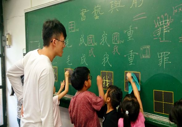 教室裡，一位男性教師和三位學生站在綠色黑板前。學生們正在用粉筆書寫中文字和幾何圖形，教師在一旁指導。黑板上方有多個中文字和數學相關的圖案。