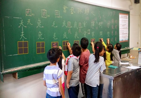 教室內，六位學生站在綠色黑板前，舉起手臂參與互動教學活動。黑板上書寫著中文字詞和方框圖示，展示漢字結構和筆畫練習內容。