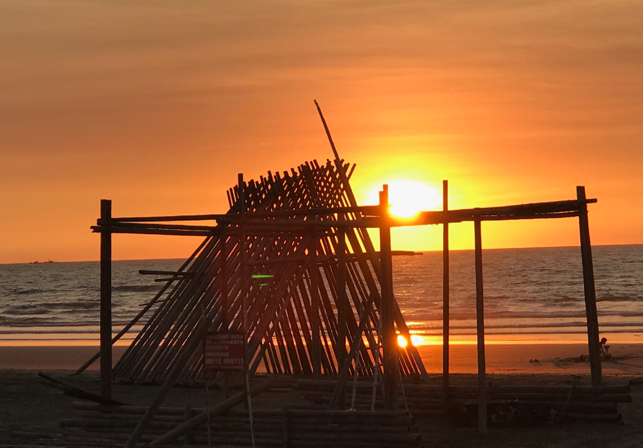 海邊沙灘上，夕陽西下，一艘廢棄船隻的木製骨架在金黃色陽光的逆光下清晰可見。船骸由多根木桿和木板組成，呈現破敗的船體輪廓，周圍豎立著木製支架。遠方水平線上波光粼粼，整個場景充滿蒼涼而詩意的氛圍。