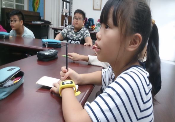 教室內，一名穿著條紋上衣的女學生坐在桌前，拿著筷子進行夾豆子活動。身後還有兩名男學生參與同樣的練習。桌上放有豆子、筷子和其他學習用具，呈現互動式教學場景。