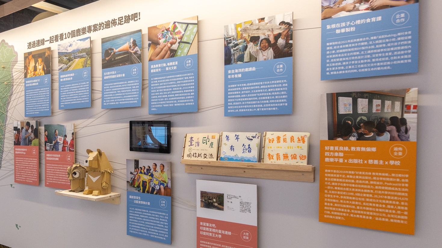 牆上展示多個藍色和橙色資訊牌，記錄教育計畫的成果與故事。展示包括十個鹿谷景點介紹、企業合作案例、學生心得分享及教育無償鄉的四方串聯等內容。木製展示架上放置標語木板和相關文宣。牆角有木製貓頭鷹雕像裝飾。