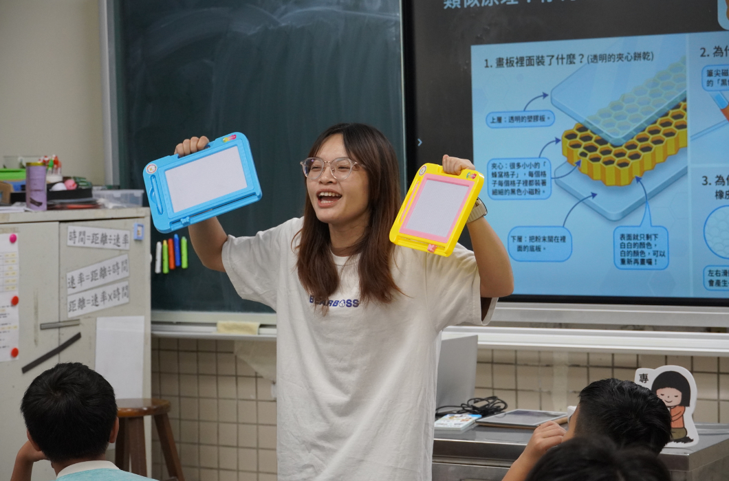 為了讓孩子更容易理解，講師雙手高高舉起兩塊彩色塑料磁性畫板（左手藍色、右手黃色），一邊對比一邊向孩子們解說。她後方的數位螢幕上正呈現「夾心餅乾」般的蜂巢格子微觀構造圖，兩者呼應，教學方式直觀且具趣味性。