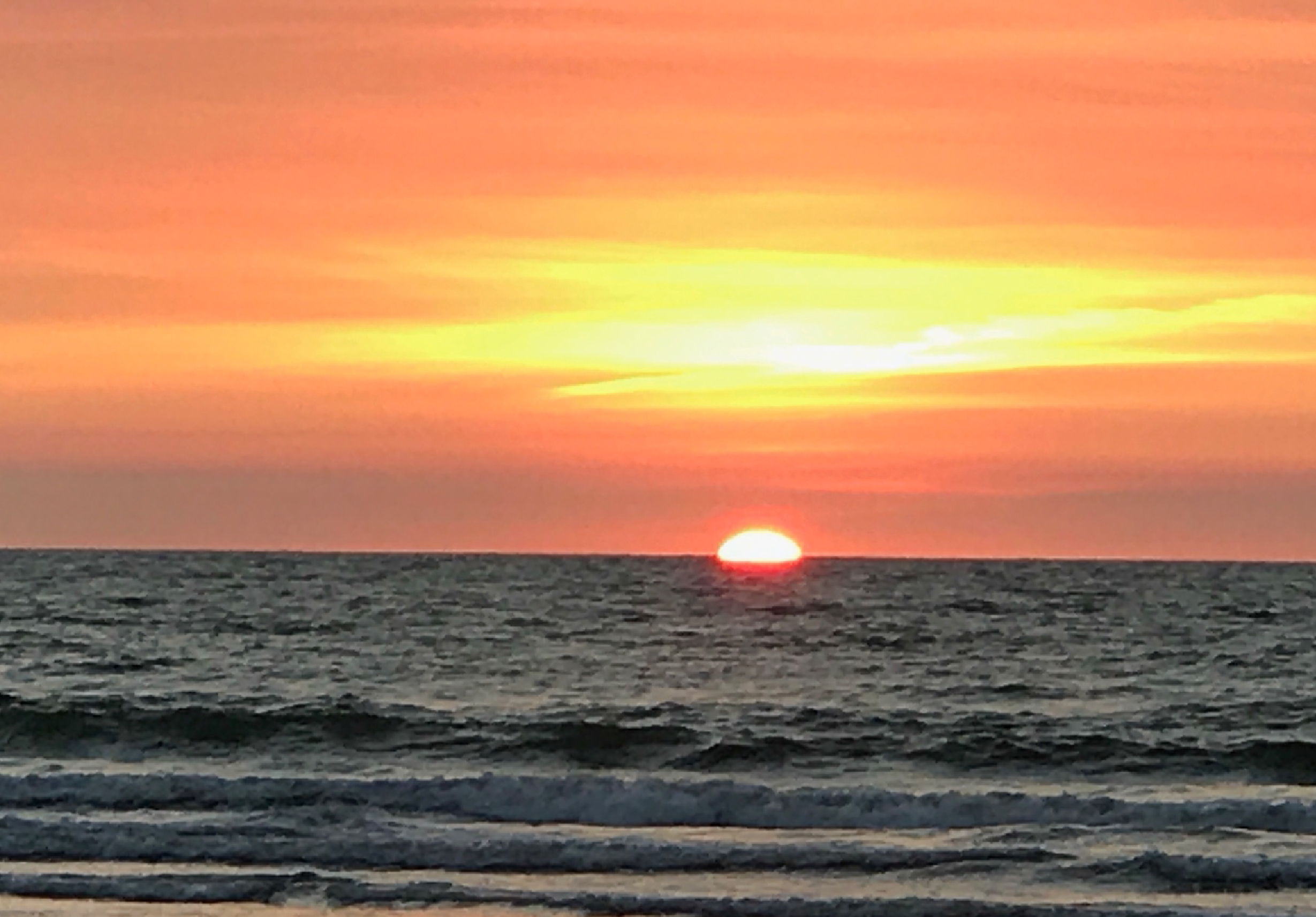 海邊日落景象，太陽半沉入地平線，天空呈現橙黃色漸層，海浪拍打沙灘，營造寧靜祥和的傍晚氛圍。