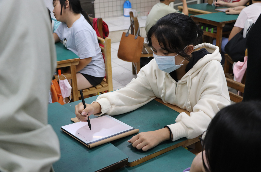 一位穿著米白色連帽上衣、戴著藍色醫用口罩的女同學獨自坐在課桌前。她左手按住桌子，右手握著黑色觸控筆，全神貫注地在平放的電子書閱讀器螢幕上進行手寫或作畫，神態極為認真。