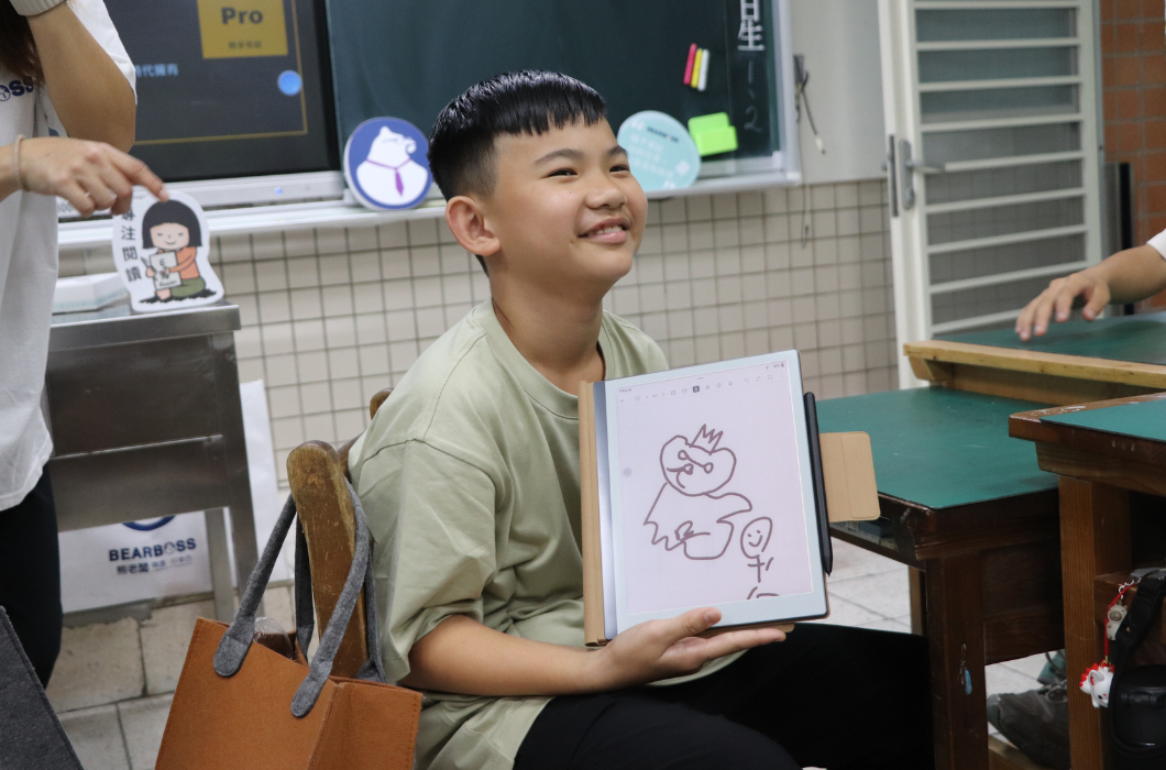 一位穿著淡綠色上衣的短髮男同學坐在椅子上，雙手托著電子書閱讀器向大家展示。螢幕上是他親手繪製的一隻戴著皇冠的熊以及一個火柴人。男孩微微揚起下巴、看著遠方，臉上帶著滿足且開心的自信微笑。