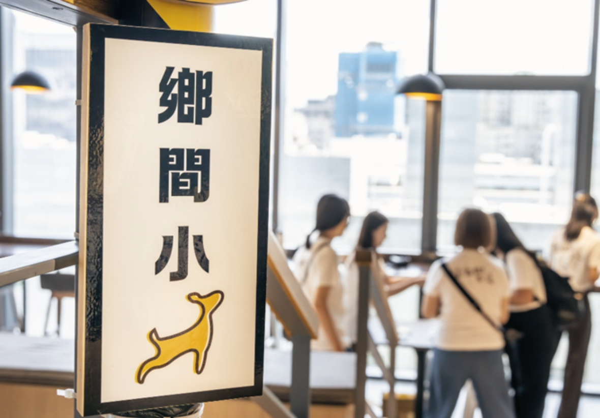 此圖為一室內場景，前景左側是一個垂直的路牌，白底上直書黑色中文字「鄉間小鹿」，下方繪有一隻黃色的梅花鹿插畫，招牌有金屬邊框。背景是一個明亮的現代開放式空間，有落地大窗，窗外可見模糊的城市建築和天際線。