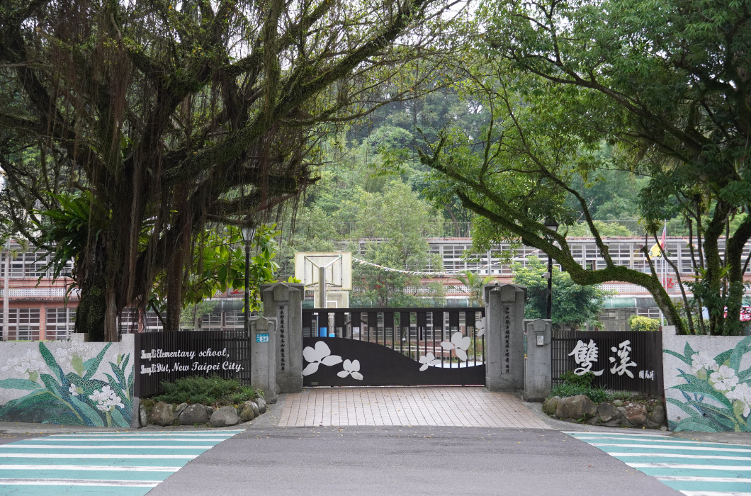 畫面為新北市雙溪國小的校門外觀。兩側石柱與白色矮牆上裝飾著優雅的綠葉與白花馬賽克磁磚拼貼，右側牆面寫著醒目的「雙溪」二字。大門上方被茂密翠綠的百年老樹枝葉溫柔覆蓋，遠處隱約可見校舍，整體氛圍寧靜且貼近自然。