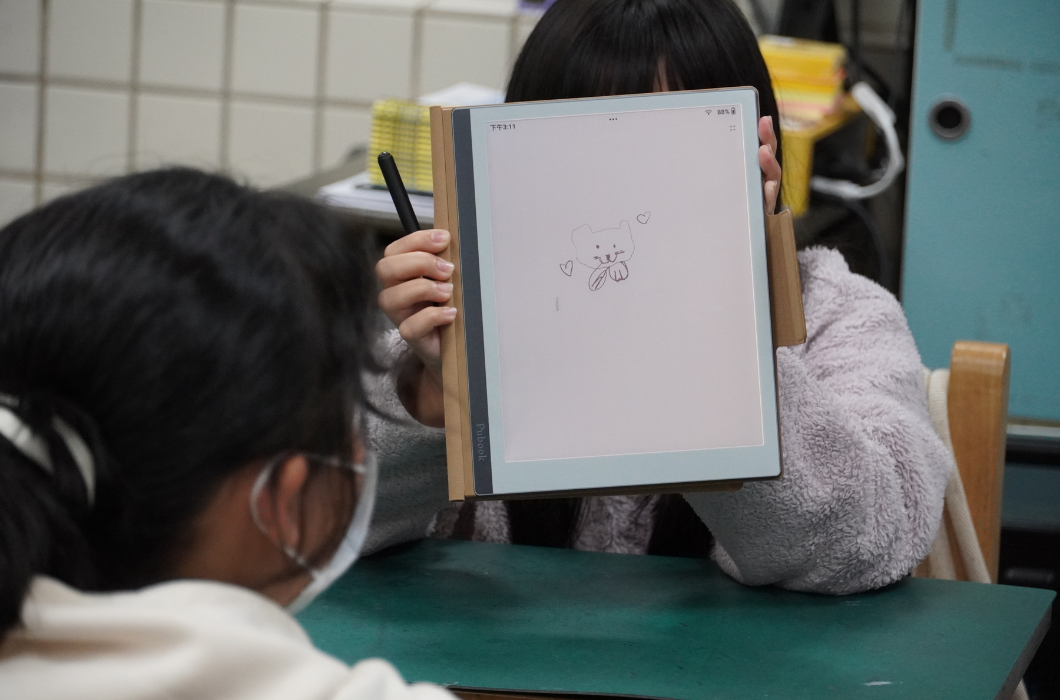 一位穿著毛絨外套的女孩坐在課桌前，將電子書閱讀器高高舉起擋在臉部前方，向對面的同學展示。螢幕正中央畫著一隻非常精緻、綁著領巾的小貓咪，兩旁還點綴著愛心，展現出電子筆細緻的線條表現。