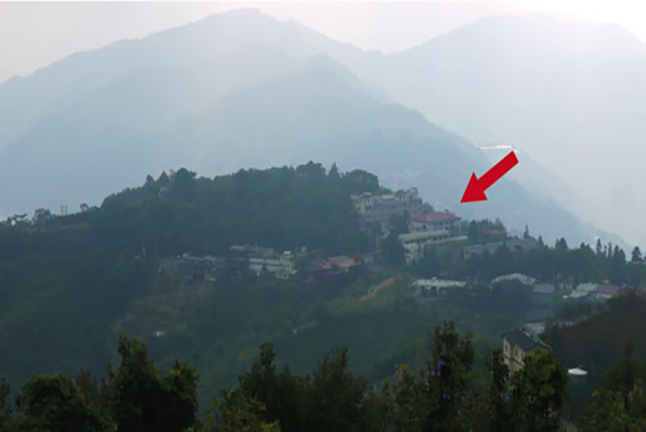 山區風景照片，遠處層疊的青翠山脈籠罩在薄霧中，山腰上散落著紅色屋頂的建築物和房舍，前景為深綠色的樹林，紅色箭頭指向右方山坡上的一棟建築。