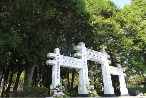 白色牌樓門形紀念碑矗立在茂密綠樹環繞的公園中，牌樓上刻有繁體中文字，門前放置紫色和白色鮮花，莊嚴肅穆的紀念設施。