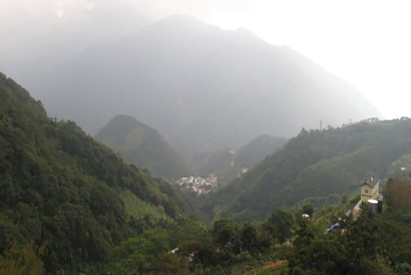 雲霧籠罩的山谷景象，兩側山坡被茂密的綠色林木覆蓋，谷底散布著白色建築物的小村莊，遠處山峰在薄霧中若隱若現，呈現寧靜的高山風景。