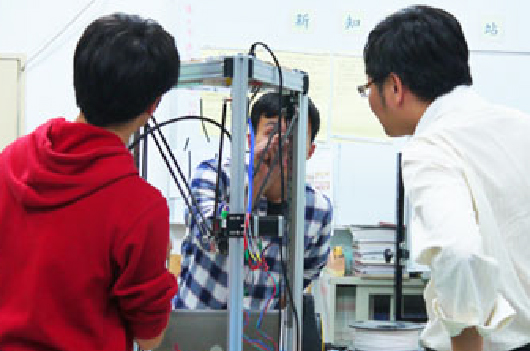 兩名學生穿著紅色和白色衣服，在實驗室中觀察和操作一台3D打印機。機器前方展示著正在列印的藍色物體。白板上方可見中文字標記，背景為實驗室環境。