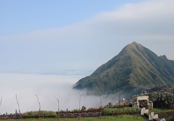 一座圓錐形火山矗立於晨霧中，山腰被白色雲霧籠罩。山下是綠色農田和稀疏的樹木，右側可見村落建築。天空呈淡藍色，雲層輕薄。