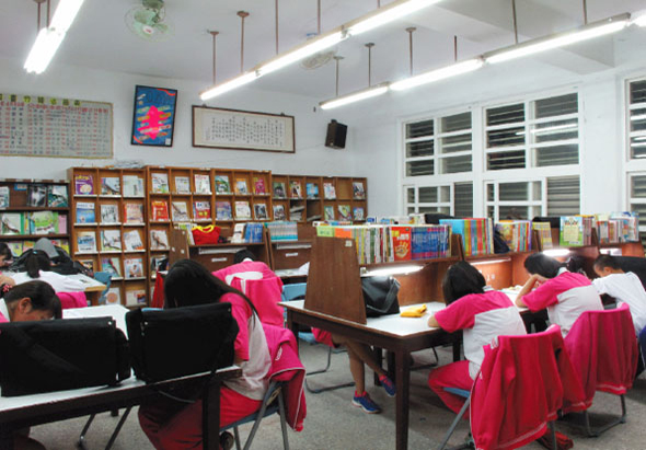 圖書館內，穿著紅色制服的學生坐在桌前閱讀。室內有書架、鋼琴、黑板和電子顯示屏。天花板裝有日光燈和通風口，窗戶採光充足。學生專注於學習，展現校園閱讀環境。