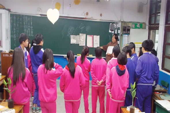 教室內，穿著粉紅色和紫色運動服的小學生們圍成一圈，面向黑板和兩位教師。室內懸掛著心形裝飾，學生們參與集體活動或課程互動。