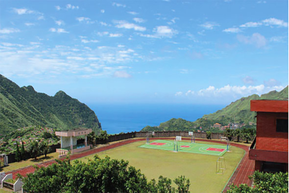 山間運動場俯瞰景觀，紅土跑道環繞綠色足球場，遠處是翠綠山巒和蔚藍海洋。場邊紅色建築物和看台清晰可見，晴空萬里，雲朵點綴。