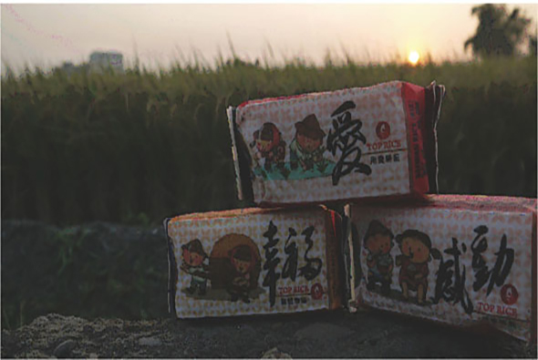 黃昏時刻，田間堆放三個紅白相間的紙箱，箱上印有卡通人物和中文字樣。背景為廣闊的麥田和遠處建築，夕陽西下映照整個農村景象。