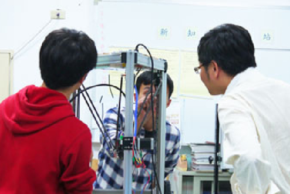 兩名學生在教室內觀察和操作3D印表機。穿紅色連帽衫的學生在左側，穿白色上衣的學生在右側，兩人正專注地檢查機器的列印頭和內部結構。背景是白色黑板。