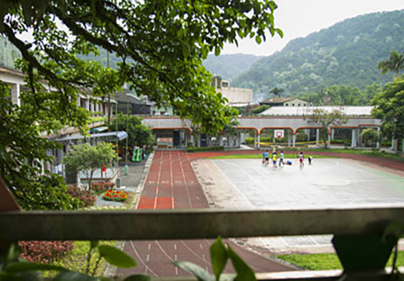 學校校園俯瞰圖，可見紅色跑道、白色籃球場、校舍建築物環繞，綠樹成蔭，背景為青山，數位學生在操場活動。