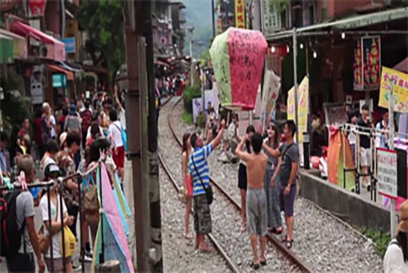 古鎮街道上舉辦節慶活動，民眾聚集在石板路上，中央有人舉著粉紅色的大型花燈。兩側建築物掛滿彩色裝飾和標語，氣氛熱鬧歡樂，呈現傳統民俗文化慶典的景象。