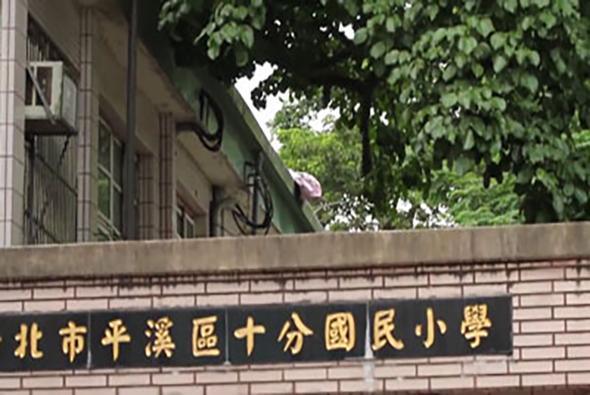 北市平溪區十分國民小學的校名牌，以黑色底板搭配金色繁體中文字，安裝在校舍前的磚牆上。背景可見校舍建築、綠色植物和樹木。