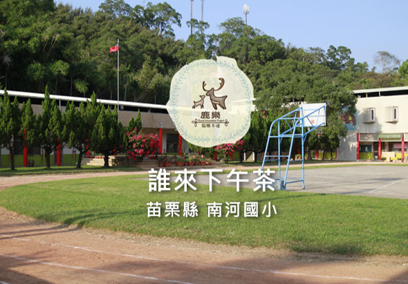 苗栗縣南河國小校園，大草坪前豎立著「誰來下午茶」活動宣傳牌，牌面以鹿的圖案為主視覺。校舍、遊樂設施與綠樹環繞，晴朗天氣。