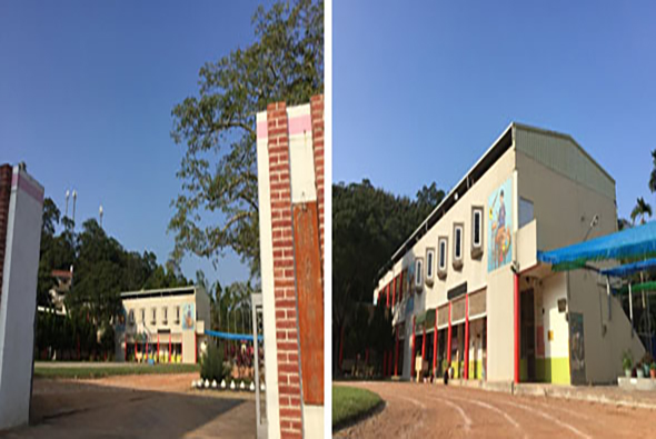 校園建築群全景，右側為主要教學樓，具有現代設計風格，外牆淺色搭配彩色門窗。左側為磚紅色立柱標誌牌。校園內綠樹環繞，地面為紅褐色路面，晴朗藍天，呈現整潔明亮的教育機構環境。