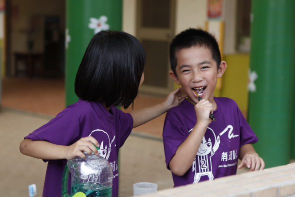 兩名穿著紫色制服的小學生在室內走廊進行科學實驗活動。左邊女孩拿著裝有液體的透明容器，右邊男孩開心地笑著，桌上放置著實驗用品。背景可見綠色柱子和教室環境。