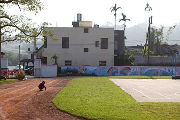 學校運動場景，一名學生在紅土跑道上運動，綠色草坪和白色籃球場在右側。背景是灰色多層建築、藍色圍欄、棕櫚樹及遠處山景。