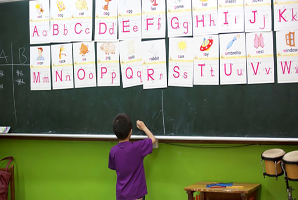 小學教室內，穿著紫色上衣的學生站在黑板前，舉手寫字。黑板上方貼滿英文字母卡片，展示大小寫A到K的字母及對應圖片。教室角落放置樂器，牆面為綠色。
