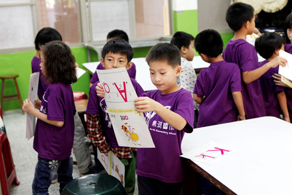 一間教室裡，穿著紫色制服的小學生們在展示他們的美術作品。中央一名男童舉著自己的繪畫作品，上面畫有紅色圖案。周圍其他學生也在觀看和分享各自的創作，背景是綠色牆面和教室佈置。