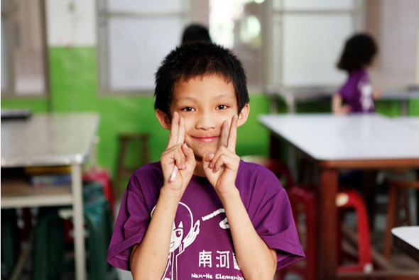 一名穿著紫色短袖上衣的小男孩在教室內擺出勝利手勢，面帶笑容看向鏡頭。背景是綠色牆面的教室環境，還有其他學生坐在桌子旁。