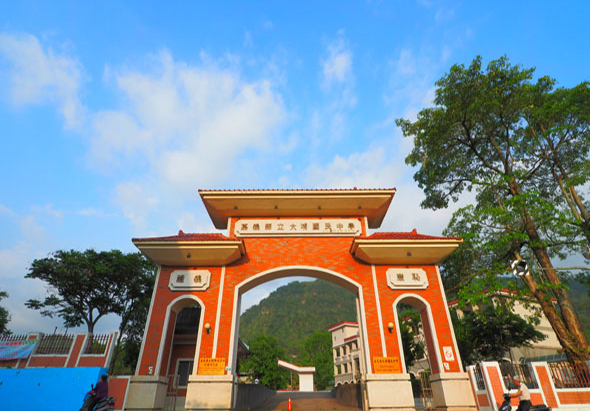 一座橙紅色傳統牌樓建築，門楣上書有中文字樣，兩側有白色窗戶，頂部為金色飾邊。背景可見綠色山脈、蒼天大樹與藍天白雲，右側有大型樟樹。這是一處具有中式建築風格的景區入口。