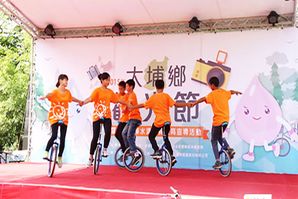 五位穿著橙色衣服的兒童在舞台上騎著單輪車表演，背景為大橋鄉社區活動的宣傳布景，舞台鋪著紅色地毯，展示特技表演。