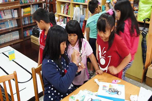 圖書館內，多名學生圍坐在桌邊進行手工藝活動。穿著深藍色波點上衣的女學生專注地操作手中的物品，其他學生在旁觀看和協助。背景可見書架、椅子和其他學生。