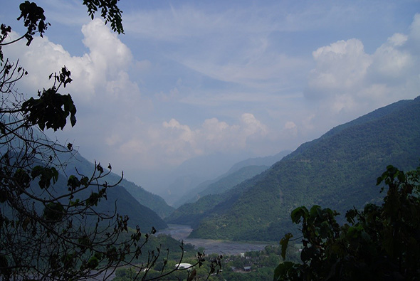 山谷風景照，遠處群山環繞，山谷中有河流流經，綠色植被覆蓋山坡，山間籠罩著薄霧，前景可見樹枝和葉片，天空飄浮著白雲，整體呈現宁靜的高山峽谷風貌。