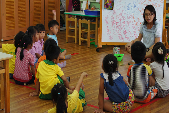 幼兒園教室內，一位女老師站在白板前進行教學，約十位穿著彩色衣服的幼童坐在木質地板上圍成半圓形聽課。教室內有木製傢俱、彩色教具和黃色儲物櫃。