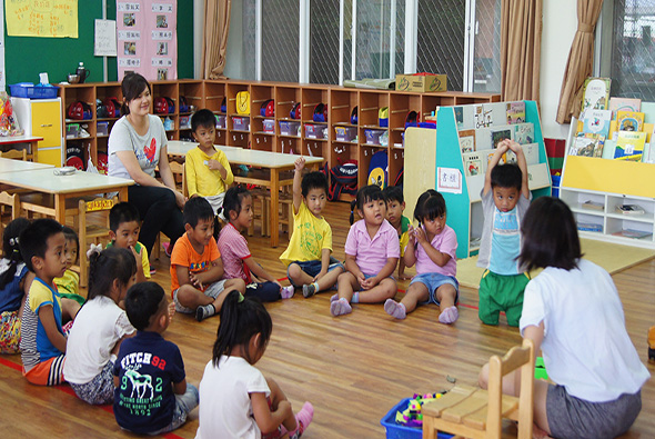 幼兒園教室內，兩名教師與約十五名穿著黃色、粉紅色等彩色上衣的幼童圍坐成圈，坐在木質地板上進行集體活動。教室背景有儲物櫃、黑板、窗戶和教學用具，營造溫暖的學習環境。