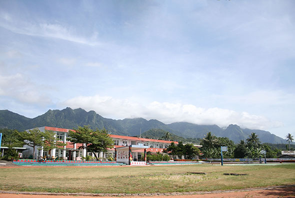 一所位於山區的小學校園，建築物為橙紅色屋頂，坐落在寬闊的紅土運動場前。背景是層疊的青綠山脈，晴朗的天空中飄浮著白雲，周圍種植著椰子樹和熱帶植被。