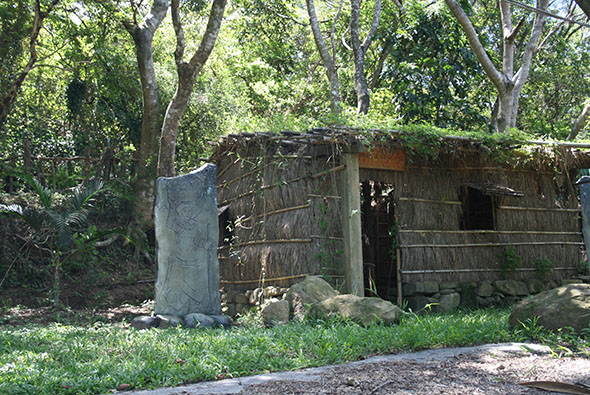 林間空地上一棟老舊木造房屋，屋頂破損、木板腐爛，門窗洞開。房屋左側立著一塊灰色石碑。周圍被大樹環繞，地面長滿草叢，呈現荒廢已久的狀態。