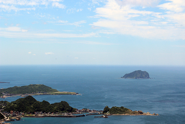 海岸線俯瞰景觀，前景為綠樹覆蓋的山丘和海灣，沿岸有房屋建築。遠方海面上可見兩座島嶼，清澈藍天與海水相映，白雲點綴其間。