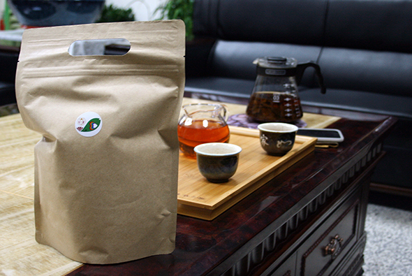 木製茶几上擺放著牛皮紙茶葉包裝袋、玻璃茶壺、黑色陶瓷茶杯和白瓷茶杯，背景為黑色沙發，展示茶葉產品與茶具的搭配場景。