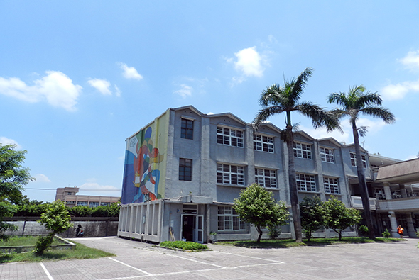 一棟灰色多層校舍建築，左側牆面繪有彩色壁畫，前方空地寬敞，種有棕櫚樹和綠樹，晴朗天氣下的學校校園景觀。