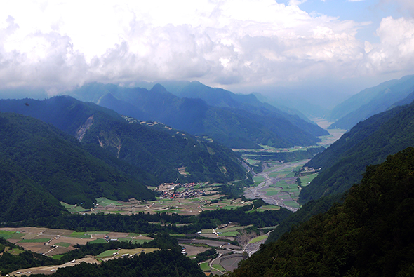 壯闊的山谷風景，翠綠的山峰環繞著寬闊的河谷平原。平原上散布著農田、村落和蜿蜒的河流，遠方山巒層疊，白雲飄浮於天空。整個景象展現出深邃的山地地形與人類聚居的和諧。