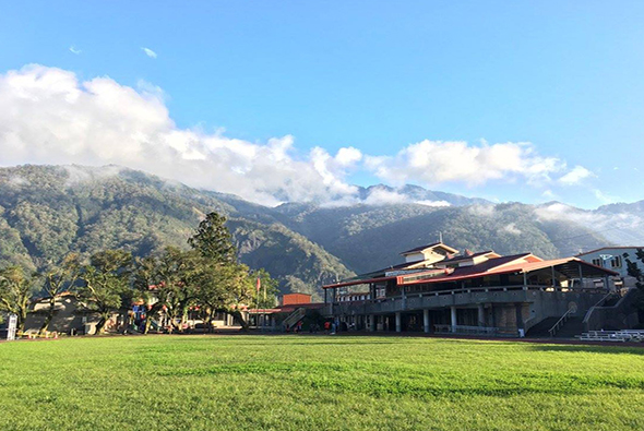 山腳下寬闊的綠色草坪，背景是雄偉的山脈和白雲。草坪邊有木造建築和散落的人群，樹木點綴其間。晴朗天空映照下，景色優美宜人。
